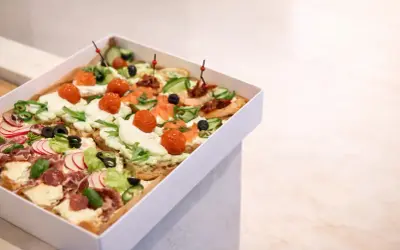 Pudełko z różnorodnymi, kolorowymi przekąskami typu bruschetta: z łososiem, pomidorkami, ogórkiem, rzodkiewką i wędliną.