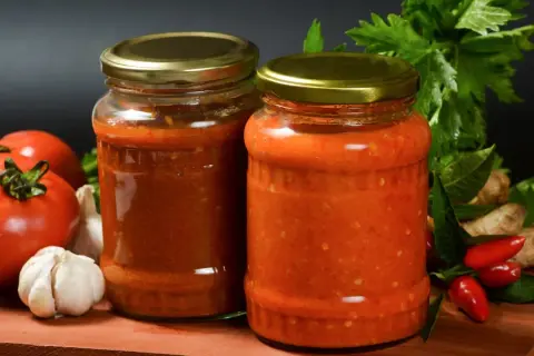 Dwa słoiki sosu pomidorowego z dodatkami: pomidory, czosnek, papryczki chili, imbir i zioła.