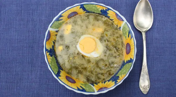 Talerz zupy szczawiowej z jajkiem i łyżka na niebieskim tle.