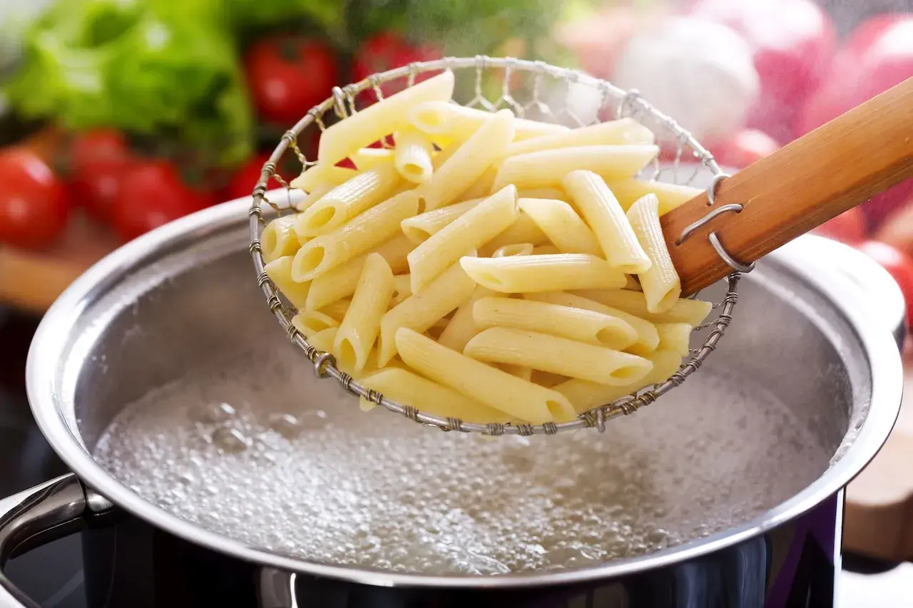 Makaron penne w cedzaku nad garnkiem z gotującą się wodą.