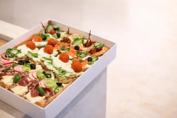Pudełko z różnorodnymi, kolorowymi przekąskami typu bruschetta: z łososiem, pomidorkami, ogórkiem, rzodkiewką i wędliną.